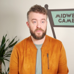 Man with a beard wearing a brown suede jacket and a green shirt, standing in front of a wall featuring the logo of Midwest Games, with indoor plants visible in the background.